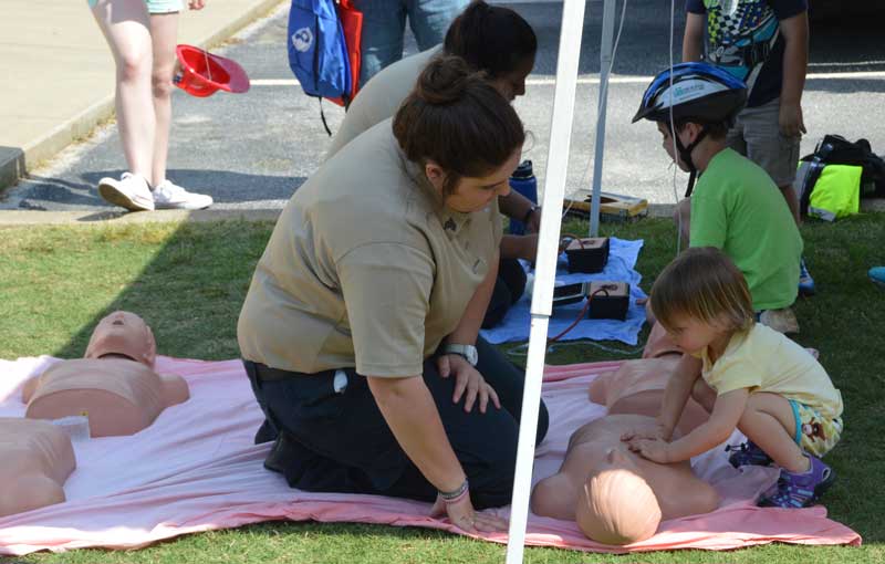 A young girl performs CPR on a rubber “dummy” as emergency medical personnel observes.